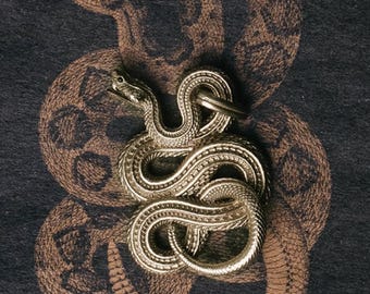 May include: A silver-colored snake pendant with intricate detailing, coiled on a dark background with a brown snake pattern. The pendant has a ring at the top for attachment. The design is detailed and textured.
