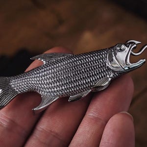 May include: A silver-colored fish-shaped bottle opener. The opener is highly detailed, with scales and fins. The fish has an open mouth with a tongue-like extension, designed to open bottles. The opener is held in a hand.