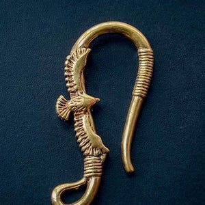 以下が含まれることがあります： 金色のイヤリングフック。鷲のデザイン。フックは湾曲しており、コイル状の部分と中央に鷲の姿があります。イヤリングは濃い青色の背景に配置されています。