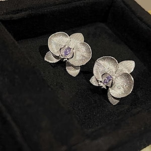 Op de afbeelding: Een paar zilverkleurige oorbellen in de vorm van orchideeën met gestructureerde, gestreepte bloemblaadjes en een enkele, kleine, peervormige paarse edelsteen in het midden van elke bloem, met gedetailleerde afwerking op het metalen oppervlak.