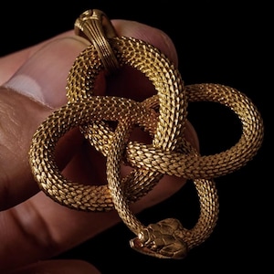 May include: A gold-coloured pendant with a snake design. The snake is highly detailed, with a textured surface and a looped, intertwined pattern. The pendant is held against a dark background.
