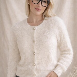 May include: A cream-colored, fuzzy, long-sleeved cardigan with a round neckline and five pearl-like buttons down the front. The garment features ribbed cuffs and a ribbed hem, crafted from a soft, textured, shaggy knit material.