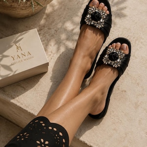 May include: Black leather sandals with a square, jeweled embellishment. The sandals feature a black rose in the center of the embellishment. The sandals are on a light-colored surface. A box with the text "M YANA ATELIER MASON" is visible.