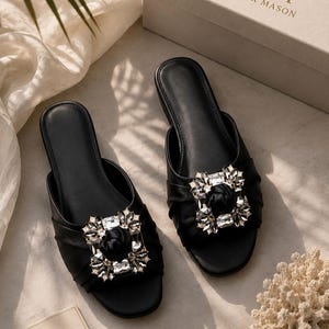 May include: Two black slip-on sandals with a large, sparkling crystal embellishment. The sandals are displayed on a neutral surface with a box and a card that reads "YANA ATELIER MASON". The sandals have a ruched design.