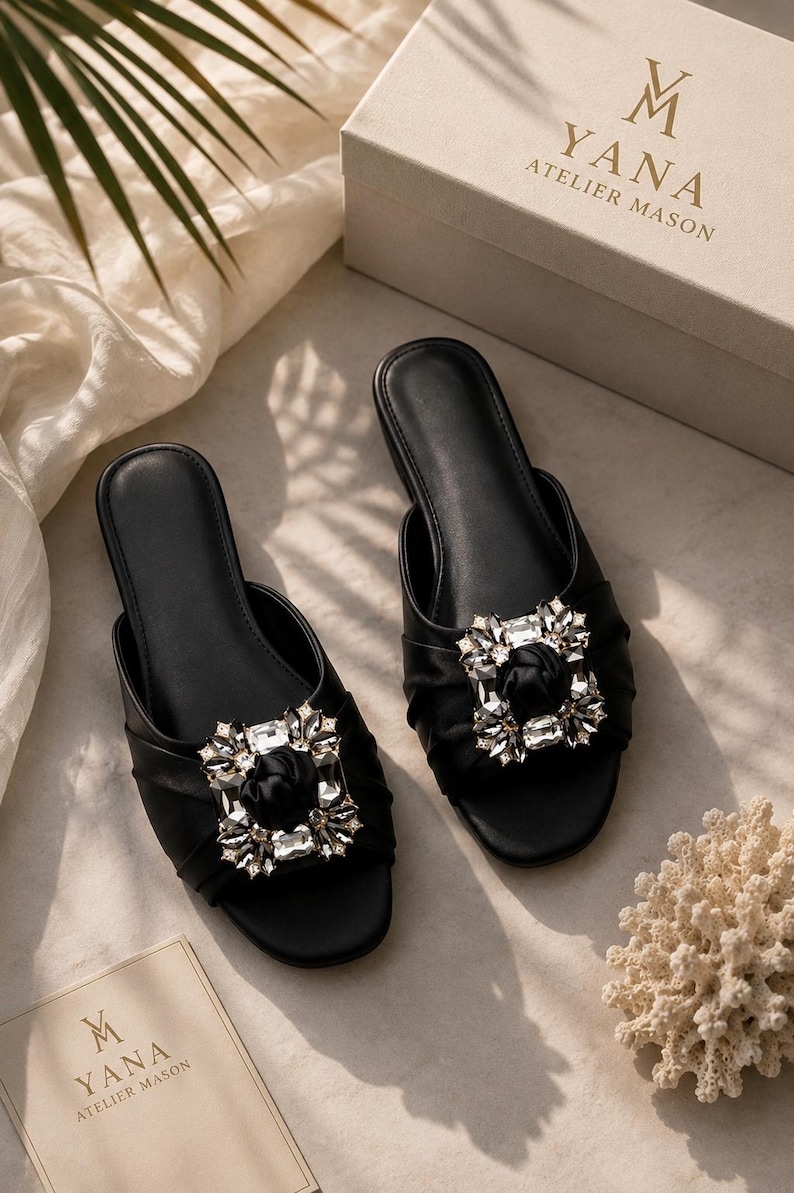 May include: Two black slip-on sandals with a large, sparkling crystal embellishment. The sandals are displayed on a neutral surface with a box and a card that reads "YANA ATELIER MASON". The sandals have a ruched design.