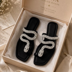 May include: Black sandals with a snake design made of sparkling rhinestones. The sandals are displayed in a box with the brand name "YANA ATELIER MASON" printed on the box and ribbon.