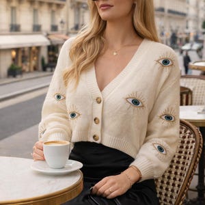 May include: Cream-colored cardigan sweater with embroidered blue eye designs. The V-neck sweater has a button-up front and is paired with a black skirt. The person is wearing black sunglasses and holding a small black purse.