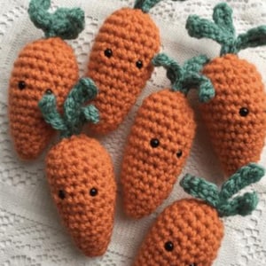 Pode incluir: Seis cenouras de crochê laranja com folhas verdes e olhos pretos. As cenouras feitas à mão estão dispostas sobre uma superfície branca texturizada, mostrando um design artesanal caprichoso.