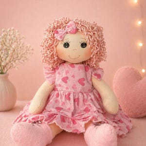 Puede incluir: Una muñeca de trapo hecha a mano con cabello rizado rosa, un lazo a juego y un vestido estampado con corazones. La muñeca tiene una cara dulce y sonriente y lleva zapatos rosas. Un corazón de peluche y un jarrón con flores blancas están en el fondo.
