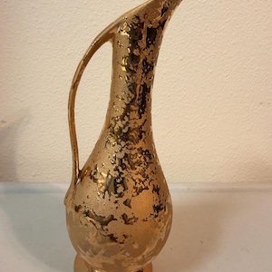 May include: A tall, slender ceramic pitcher with a metallic gold finish. The surface features a mottled, textured pattern with irregular patches of gold and lighter tones. It has a curved handle and a flared, elongated spout.