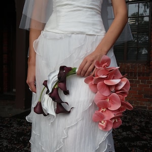 May include: A strapless white wedding dress with a layered skirt. The bride holds a bouquet of white calla lilies and dark purple flowers, along with a cascade of pink orchids. A sheer veil is draped over the bride's head.