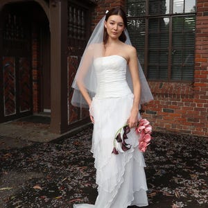 May include: A bride in a strapless white wedding dress with layered ruffles and a flowing train. She wears a sheer veil and holds a bouquet of pink and dark purple flowers. The background features a brick building and a wooden door.