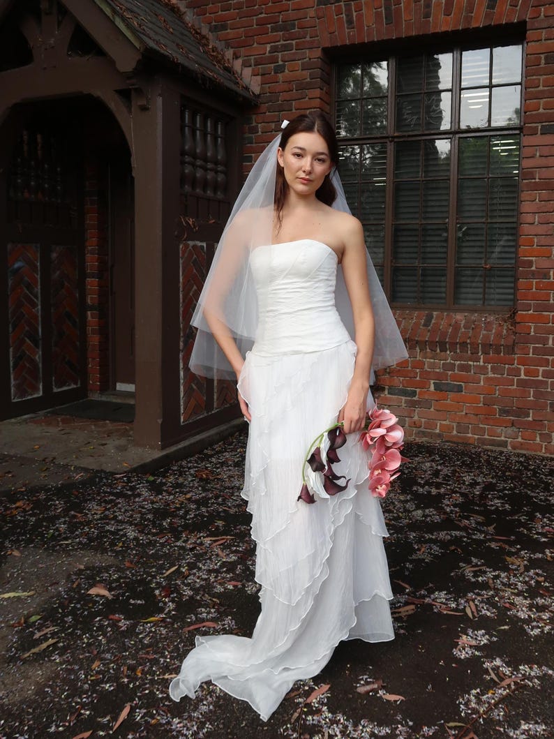 May include: A bride in a strapless white wedding dress with layered ruffles and a flowing train. She wears a sheer veil and holds a bouquet of pink and dark purple flowers. The background features a brick building and a wooden door.