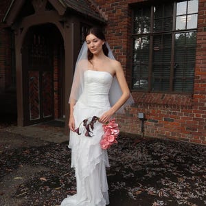 May include: A strapless white wedding dress with a layered skirt and a long train. The bride is holding a bouquet of pink flowers and dark purple calla lilies. She is wearing a sheer veil. The background is a brick building.