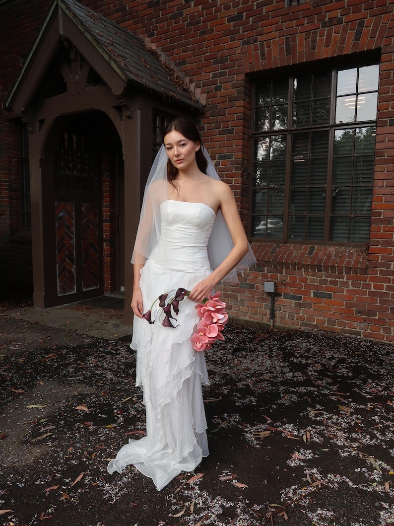 May include: A strapless white wedding dress with a layered skirt and a long train. The bride is holding a bouquet of pink flowers and dark purple calla lilies. She is wearing a sheer veil. The background is a brick building.