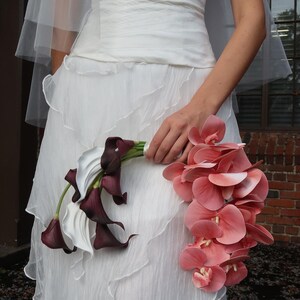 May include: A white wedding dress with a layered skirt and a sheer veil. The bride holds a bouquet of white calla lilies and deep burgundy calla lilies, along with a cascade of pink orchids.