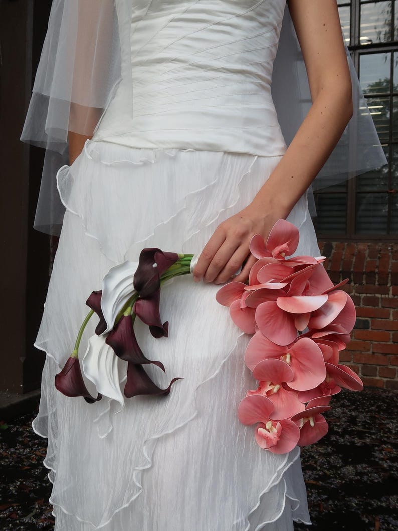May include: A white wedding dress with a layered skirt and a sheer veil. The bride holds a bouquet of white calla lilies and deep burgundy calla lilies, along with a cascade of pink orchids.