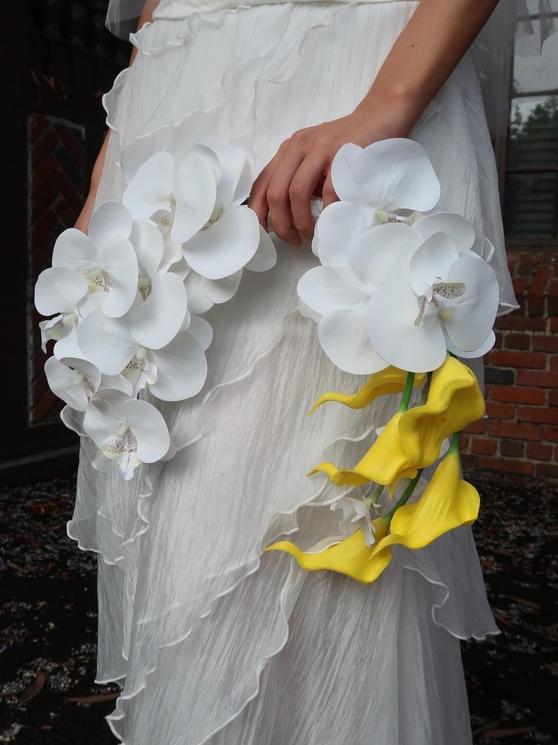 May include: A white wedding dress with a ruffled design. The dress is paired with two bouquets of flowers. One bouquet features white orchids, and the other features yellow calla lilies.