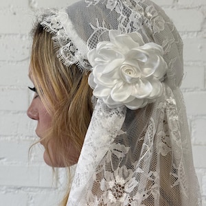 May include: A white lace veil with a floral design and a large white satin flower. The veil is draped over the head and shoulders. The lace has a delicate pattern, and the flower is a prominent accent.