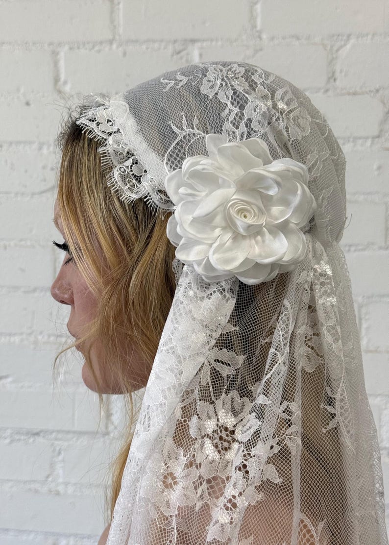May include: A white lace veil with a floral design and a large white satin flower. The veil is draped over the head and shoulders. The lace has a delicate pattern, and the flower is a prominent accent.