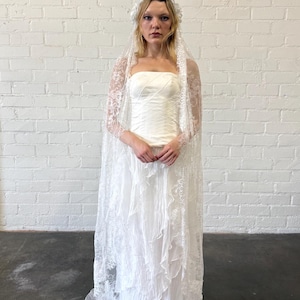 May include: A white strapless wedding dress with a layered skirt and a lace veil. The dress has a fitted bodice and a flowing skirt. The veil is long and sheer, with delicate lace detailing. The bride is wearing a floral headpiece.