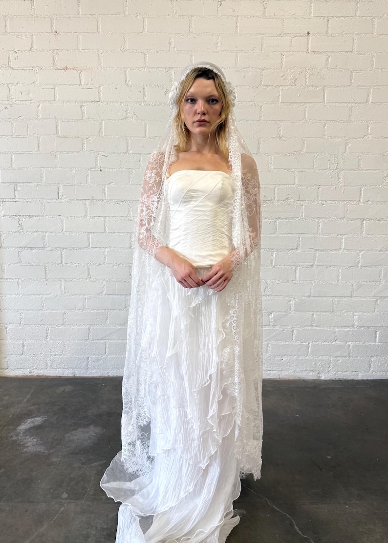 May include: A white strapless wedding dress with a layered skirt and a lace veil. The dress has a fitted bodice and a flowing skirt. The veil is long and sheer, with delicate lace detailing. The bride is wearing a floral headpiece.
