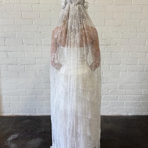 May include: A white wedding dress with a long, flowing skirt and a lace overlay. The dress is paired with a matching lace veil, adorned with floral accents. The bride is standing in front of a white brick wall.