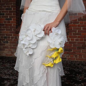 May include: A white wedding dress with a layered skirt and a bodice. The dress is adorned with white orchids and yellow calla lilies. The bride is holding the flowers.