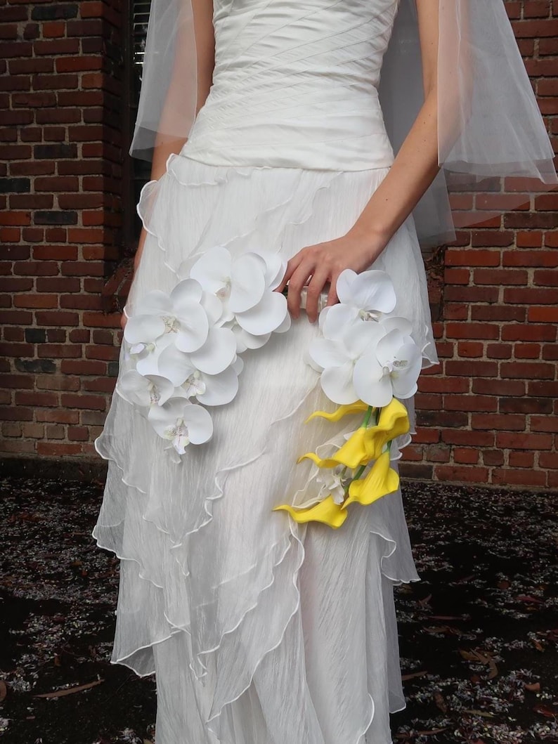 May include: A white wedding dress with a layered skirt and a bodice. The dress is adorned with white orchids and yellow calla lilies. The bride is holding the flowers.