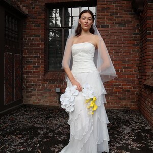 May include: A strapless white wedding dress with layered ruffles and a flowing train. The bride is holding a bouquet of white and yellow flowers. A sheer veil is draped over the head. The background is a brick building.