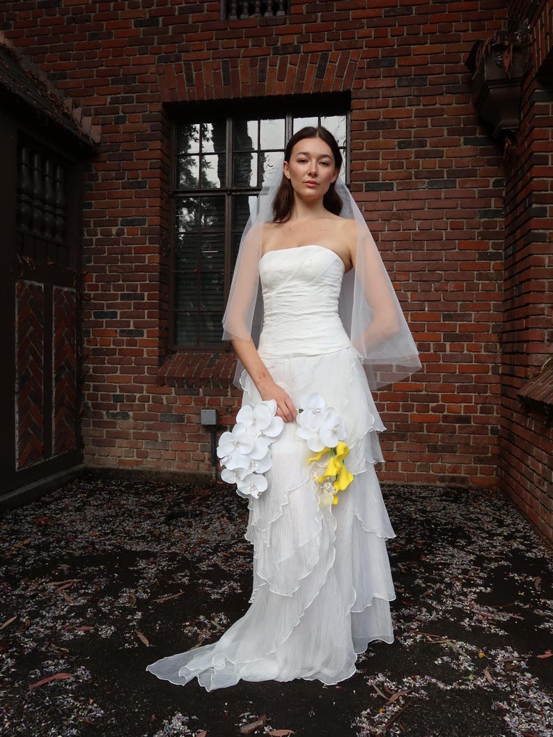 May include: A strapless white wedding dress with layered ruffles and a flowing train. The bride is holding a bouquet of white and yellow flowers. A sheer veil is draped over the head. The background is a brick building.