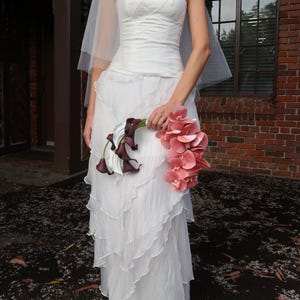 May include: A strapless white wedding dress with a layered skirt and a long train. The bride is holding a bouquet of pink orchids and dark purple calla lilies. A sheer veil is draped over the head.