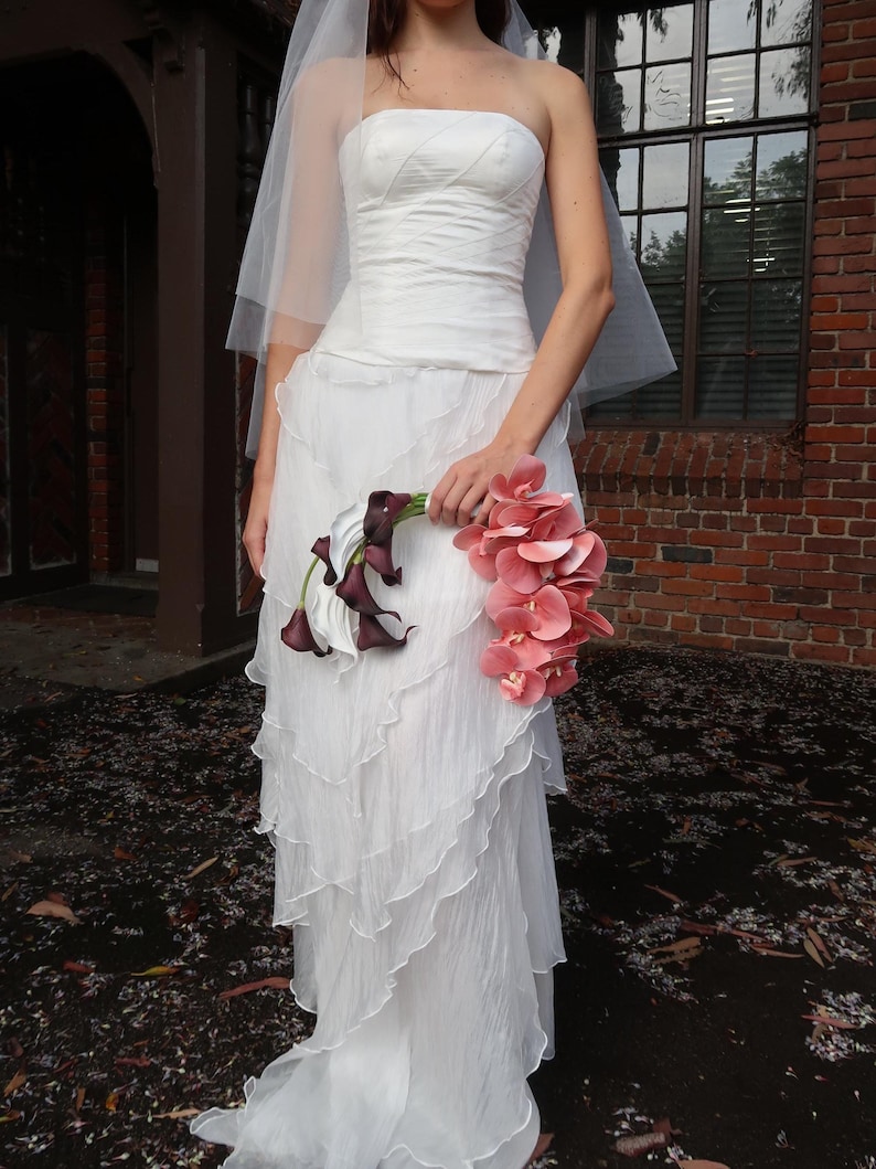 May include: A strapless white wedding dress with a layered skirt and a long train. The bride is holding a bouquet of pink orchids and dark purple calla lilies. A sheer veil is draped over the head.