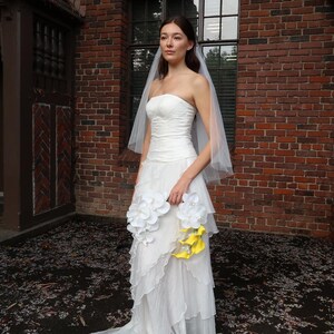 May include: A strapless white wedding dress with a layered skirt and a long train. The bride is holding a bouquet of white and yellow flowers. She is wearing a sheer veil. The background is a brick building.