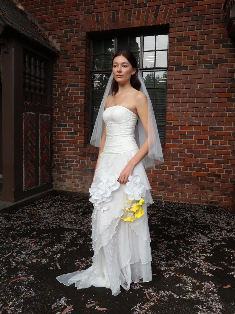 May include: A strapless white wedding dress with a layered skirt and a long train. The bride is holding a bouquet of white and yellow flowers. She is wearing a sheer veil. The background is a brick building.