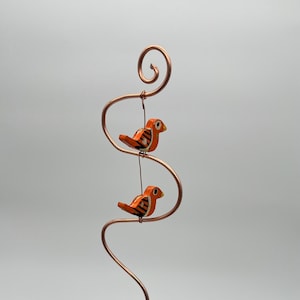 May include: A copper wire sculpture with two small, orange and black bird figurines. The birds are suspended from the wire, which is shaped into a decorative, flowing design. The sculpture is held by a hand against a plain background.