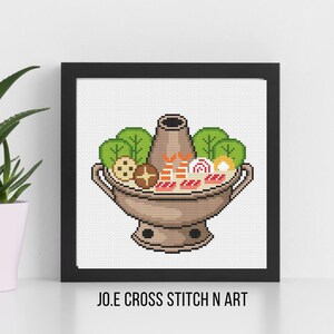 May include: A framed cross-stitch artwork featuring a tan-coloured hot pot filled with green leafy vegetables, orange shrimp, pink-swirled fish cakes, and brown mushrooms. The text JO.E CROSS STITCH N ART is printed in black below the frame.