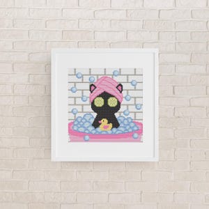 May include: A framed cross-stitch pattern featuring a black cat in a pink bathtub filled with blue bubbles. The cat wears a pink towel wrap on its head and two green cucumber slices over its eyes. A small yellow rubber duck sits in the bubbles.