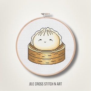 May include: A cross-stitch embroidery in a circular wooden hoop featuring a white dumpling with a smiling face sitting inside a brown bamboo steamer basket. The text JO.E CROSS STITCH N ART is printed in black capital letters below the hoop.