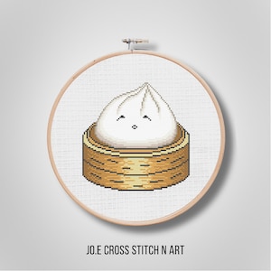 May include: Cross-stitch embroidery of a cartoon dumpling in a bamboo steamer basket. The dumpling is white with a smiling face. The basket is brown and tan. The embroidery is set in a wooden hoop. Text at the bottom reads "JO.E CROSS STITCH N ART".