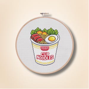 May include: Cross-stitch embroidery of a cup of noodles in a wooden hoop. The design features a cup with the words "YUMMY CUP NOODLES" and a pixelated image of noodles, meat, an egg, and vegetables.