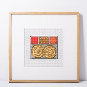 May include: Cross-stitch art of a meal, featuring two spiral-shaped items and three side dishes in a rectangular tray. The side dishes are red, brown, and orange. The artwork is framed in a light wood frame.