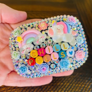 May include: A rectangular case covered in iridescent rhinestones, featuring a white unicorn, a pastel rainbow, and small resin roses. The text CHILL PILLS is spelled out using colorful round beads with gold letters, accompanied by a blue heart bead.