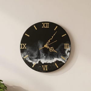 May include: A circular black wall clock with gold Roman numerals and hands. The clock face has a white and grey wave design. The clock is mounted on a light beige wall.