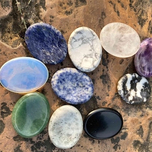 May include: An assortment of oval-shaped polished gemstones in various colors, including blue, white, green, and black. The stones are arranged on a textured, brown surface, showcasing their unique patterns and textures.