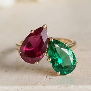 May include: A gold ring featuring two pear-shaped gemstones: a deep red ruby and a vibrant green emerald. The gemstones are set side-by-side, creating a unique and eye-catching design. The ring is displayed on a neutral-toned surface, highlighting the colors of the gemstones.
