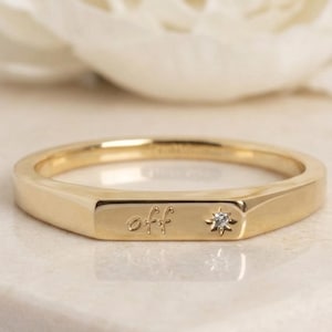 May include: A gold band ring featuring a flat rectangular front surface. The surface is engraved with the lowercase script text "off" and is accented with a small, embedded star-shaped setting containing a single clear, round gemstone.