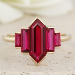 May include: A gold ring featuring a central, rectangular, deep red gemstone flanked by four smaller, rectangular red gemstones. The ring has a geometric design and is set against a neutral background.