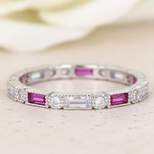 May include: A silver-toned band ring featuring an alternating pattern of rectangular-cut vibrant pink stones and round-cut clear stones, accented by a single rectangular-cut clear stone centred in the design with milgrain detailing.