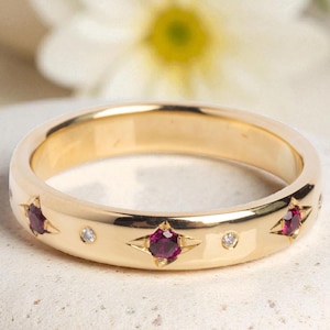 Könnte beinhalten: Ein goldener Bandring mit drei sternförmigen Fassungen, die jeweils einen tiefroten Edelstein halten. Kleine, klare Edelsteine sind zwischen den Sternen gesetzt. Der Ring liegt auf einer weißen, strukturierten Oberfläche.